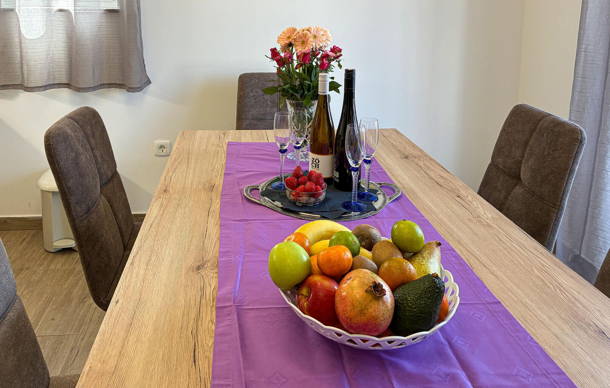 Ein Esstisch mit lila Tischdecke, einer Obstschale, einer Vase mit Blumen und Weinflaschen.