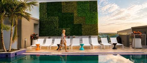 A swimming pool with lounge chairs, a woman walking, and a green wall.