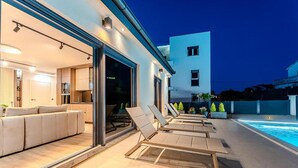 Un espacio exterior moderno con piscina, sillas de tomar el sol y una cocina visible a través de puertas de vidrio.