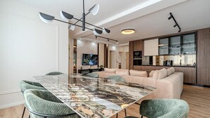 Una sala moderna con una mesa de comedor de mármol, sillas tapizadas en verde, un sofá color beige y una zona de cocina con armarios de madera y un ventanal grande.