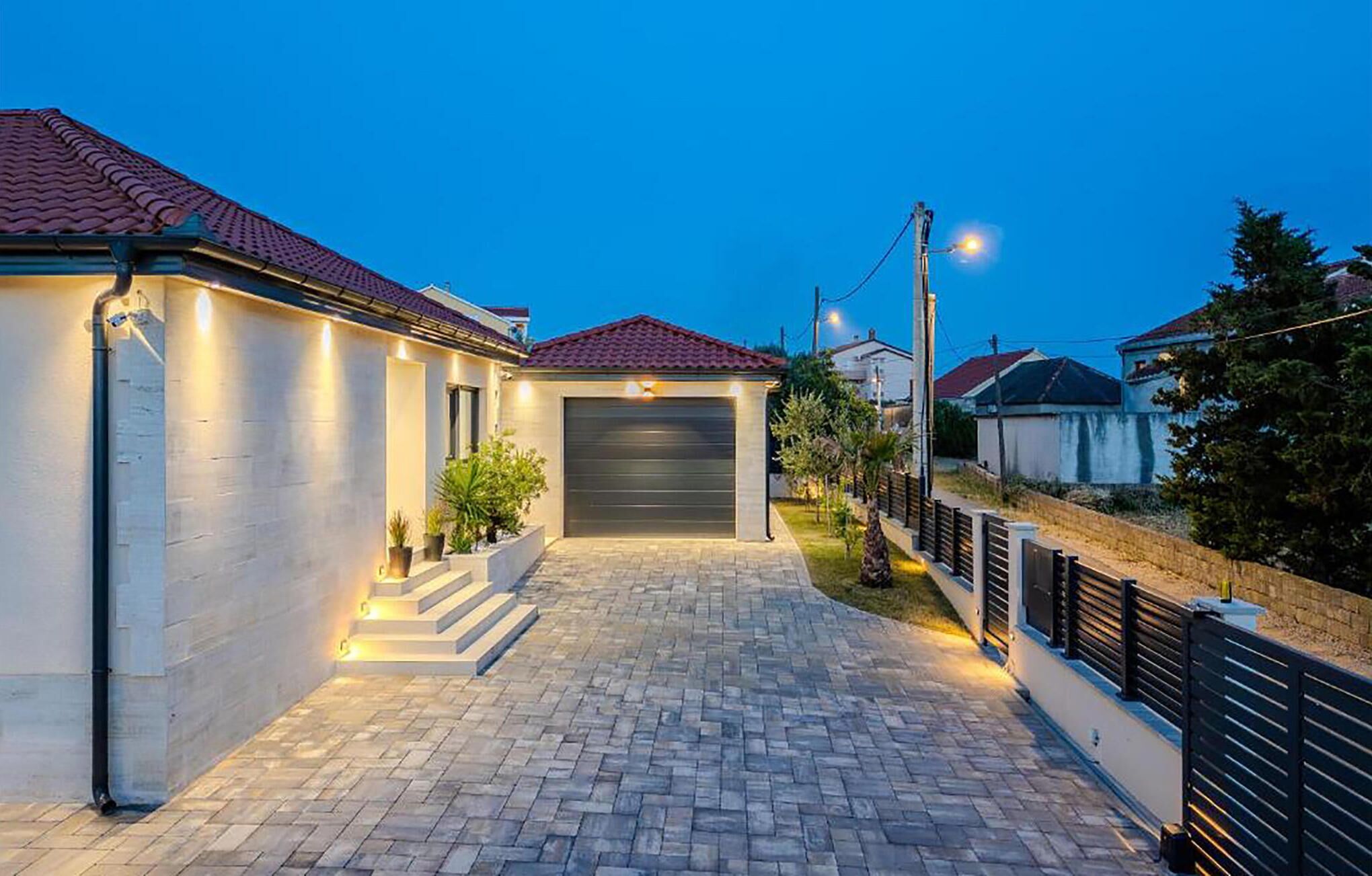 Une maison moderne avec une allée pavée menant à un garage, entourée d’une clôture basse et de verdure.