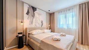 Habitación de hotel moderna con una cama grande, una mesita auxiliar pequeña, una lámpara de pared y un ventanal con cortinas.