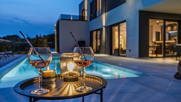 Ein zeitgemäßer Außenbereich mit Pool, zwei Gläsern Rotwein und einer Kerze auf einem Tisch.