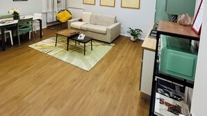 Een moderne woonkamer met een bank, salontafel en een eethoek. Er hangen afbeeldingen in lijsten aan de muur en is er een keuken met een koelkast en een magnetron.