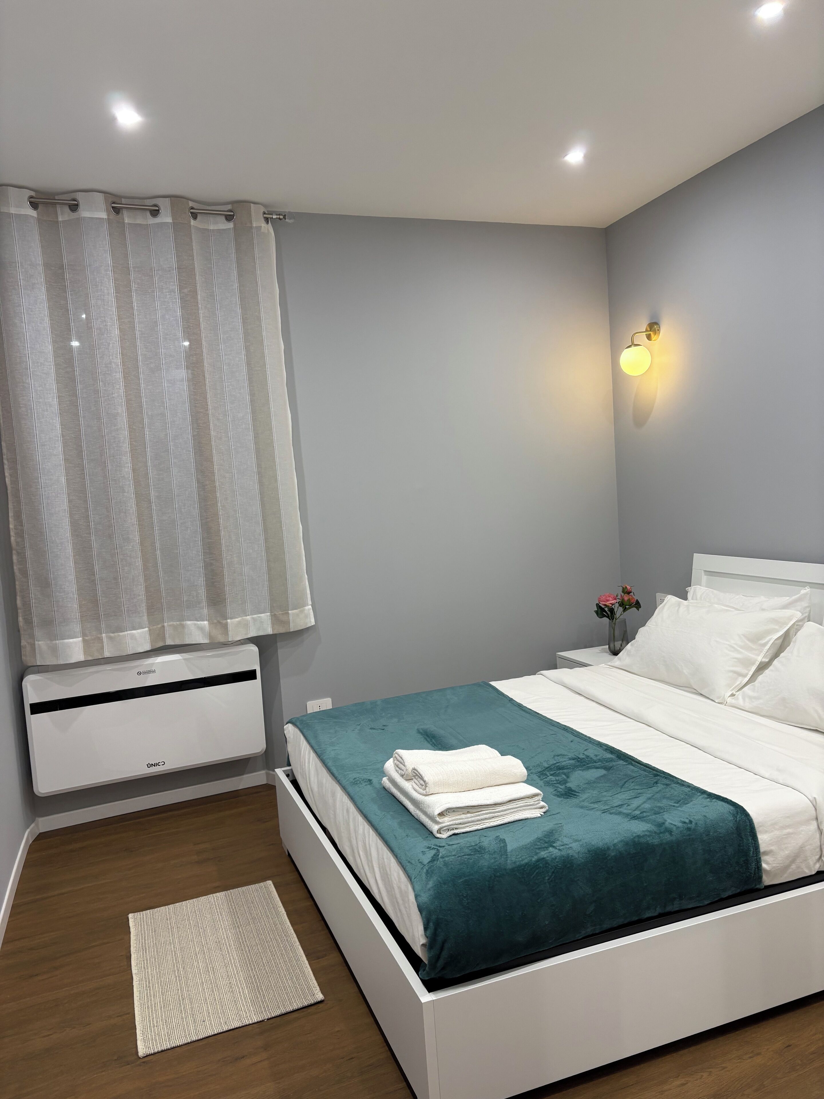 Un dormitorio con cama, una mesita de noche, una lámpara de pared, una ventana con cortinas y un aire acondicionado de pared.