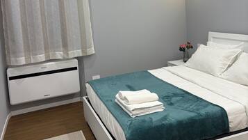 Un dormitorio con cama, una mesita de noche, una lámpara de pared, una ventana con cortinas y un aire acondicionado de pared.