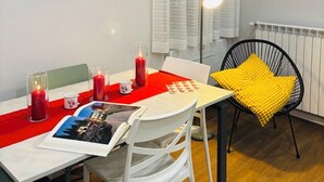 Een eethoek met een tafel gedekt voor twee personen, een lijst met een afbeelding van de scheve toren van Pisa, een rode kaars en een geel kussen op een zwarte stoel.