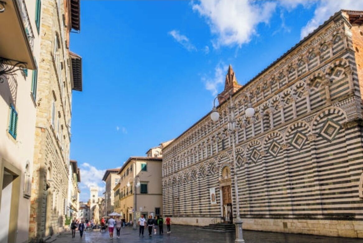 Una stretta via fiancheggiata da edifici storici, con una fontana e un cielo azzurro intenso.