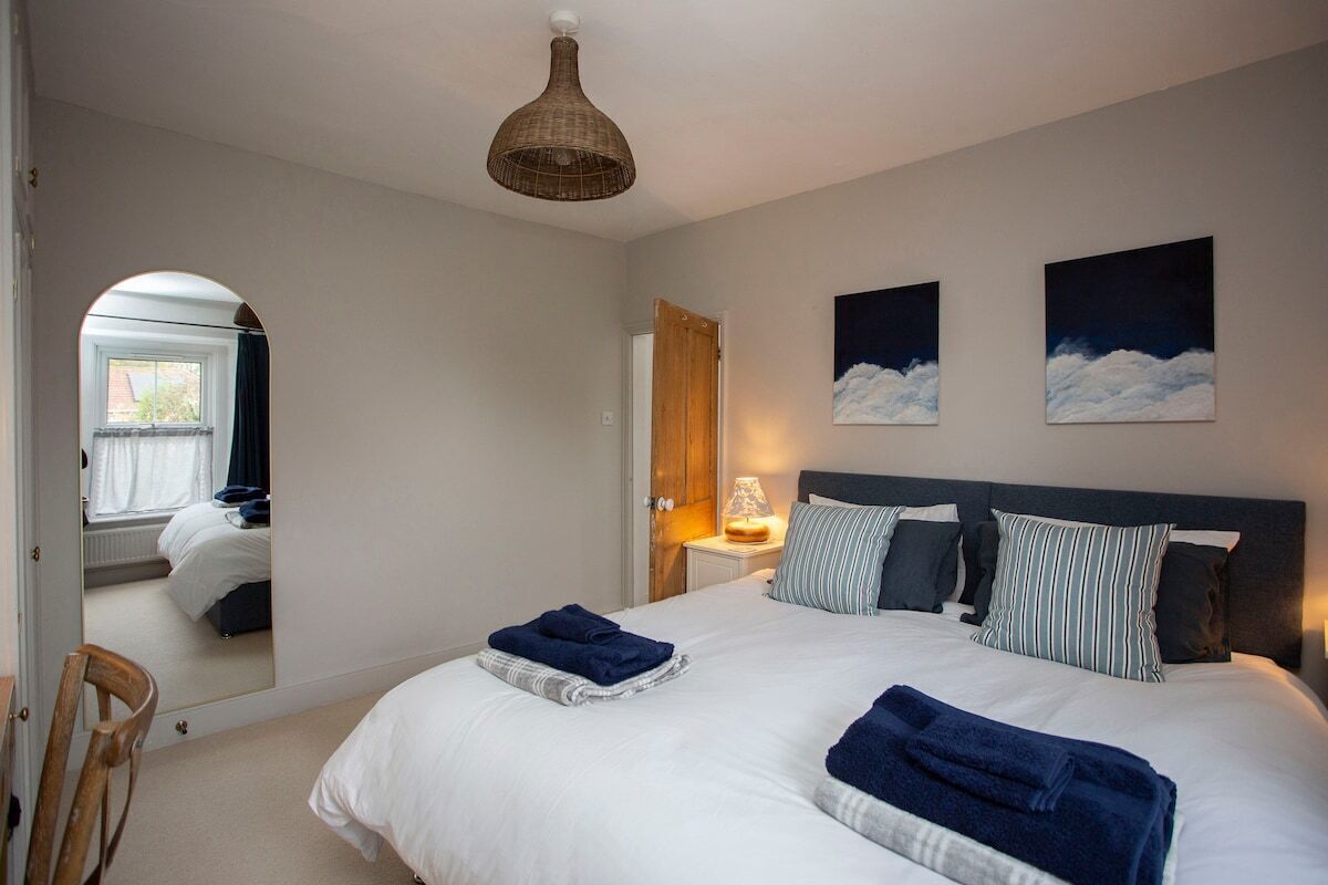 A bedroom with a large bed, a bedside table with a lamp, a mirror, and two paintings on the wall.