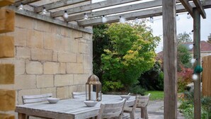 Une pergola en bois décorée de guirlandes lumineuses, un mur en pierre et un jardin agrémenté d’arbres et de fleurs.