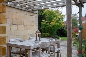 A wooden pergola with string lights, a stone wall, and a garden with trees and flowers. - Charming Family Cottage in Bath w/Parking (Bath)