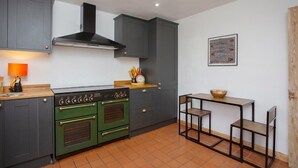 Une cuisine de taille réduite, avec des meubles de couleur sombre, un poêle vert et une petite table à manger avec des chaises.