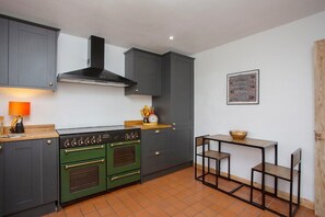 Une cuisine de taille réduite, avec des meubles de couleur sombre, un poêle vert et une petite table à manger avec des chaises.