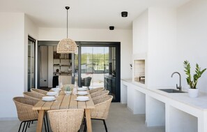 A dining area with a wooden table set for six, surrounded by wicker chairs, a woven pendant light, and a view through a glass door to a kitchen area. - Luxurious home in Nova Vas (Nova Vas)