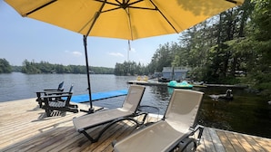 A wooden deck with two lounge chairs and a yellow umbrella, overlooking a calm lake with boats and a dock in the background. - Dock n Dive (Bala)