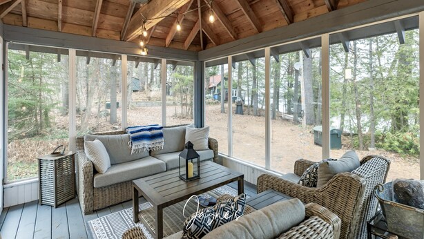 A covered porch with wicker furniture, a wooden table, and large windows offering a view of the forest. - Dock n Dive (Bala)