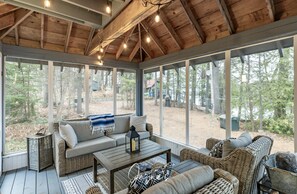 Eine überdachte Veranda mit Korbmöbeln, einem Holztisch und großen Fenstern mit Blick auf den Wald.