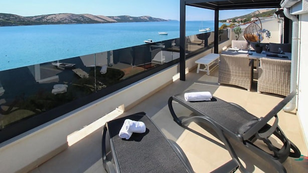A balcony with two lounge chairs, a small table, and a glass barrier overlooking a body of water with boats. - Amazing apartment in Stara Novalja with heated swimming pool (Stara Novalja)
