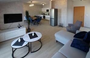 Ein modernes Wohnzimmer mit einer Couch, zwei runden Tischen, einem Fernseher und einer Küchenzeile inklusive Kühlschrank und Mikrowelle.