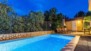A swimming pool with a stone border, surrounded by a fence and trees. There are lounge chairs and a building with a door and a light fixture. - Gorgeous apartment in Silo with outdoor swimming pool (Silo)