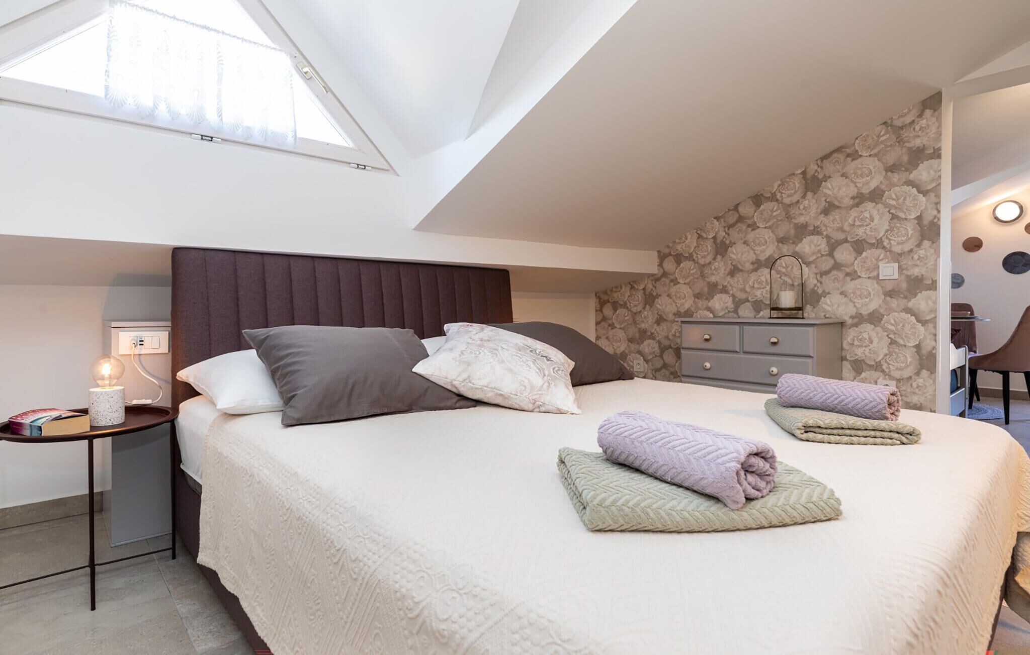 Une chambre avec un plafond en pente, un lit agrémenté de plusieurs coussins, une table de chevet avec une lampe et une commode recouverte d’un papier peint à motifs floraux.