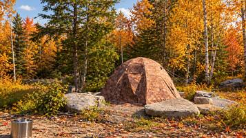 Tienda de campaña instalada en un bosque con follaje otoñal, rodeada de rocas y un contenedor metálico en el suelo.