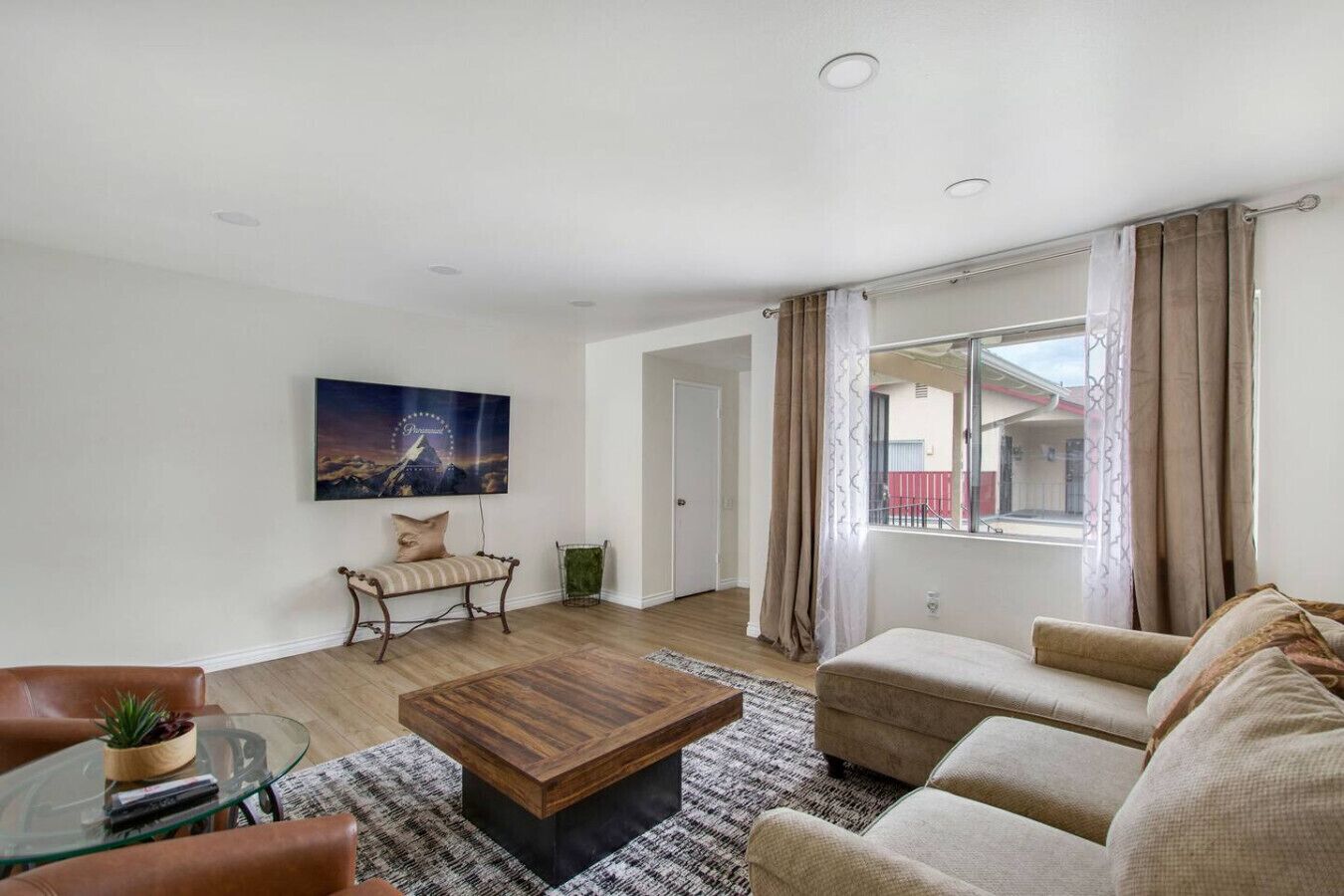 A modern living room with a wooden coffee table, a sofa, a glass table with a plant, and a large window with curtains.