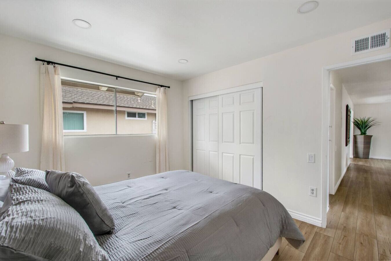 Een slaapkamer met een bed, een raam met gordijnen, een witte deur en een gang die naar een andere kamer leidt.