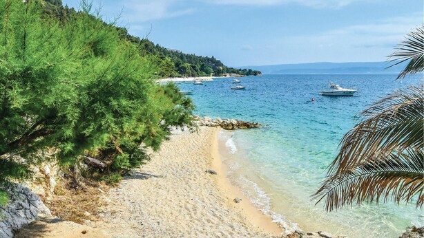 Ein Strand mit klarem türkisblauem Wasser, Booten und einem bewaldeten Hügel.