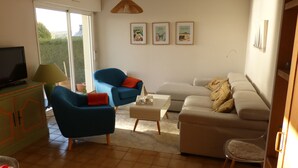 Een woonkamer met een televisie, twee fauteuils, een bank en een salontafel. Drie ingelijste foto's hangen aan de muur en er is een raam met uitzicht op groen.