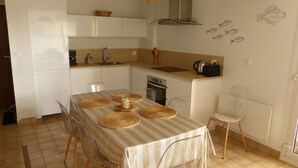 Een kleine keuken met witte kastjes, een kookplaat en een gootsteen. Er staan vier stoelen rond een tafel met geweven tafelonderleggers.