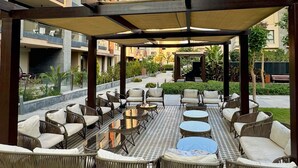An outdoor seating area with wicker chairs and tables, a checkered floor, and a covered section with a pergola. - spacious apartment w/ pool and garden view (Cairo Governorate)