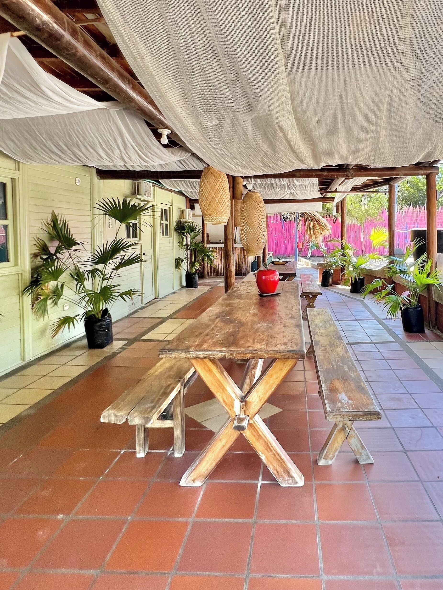 A covered outdoor dining area with a long wooden table and benches, surrounded by potted plants and a pink fence.