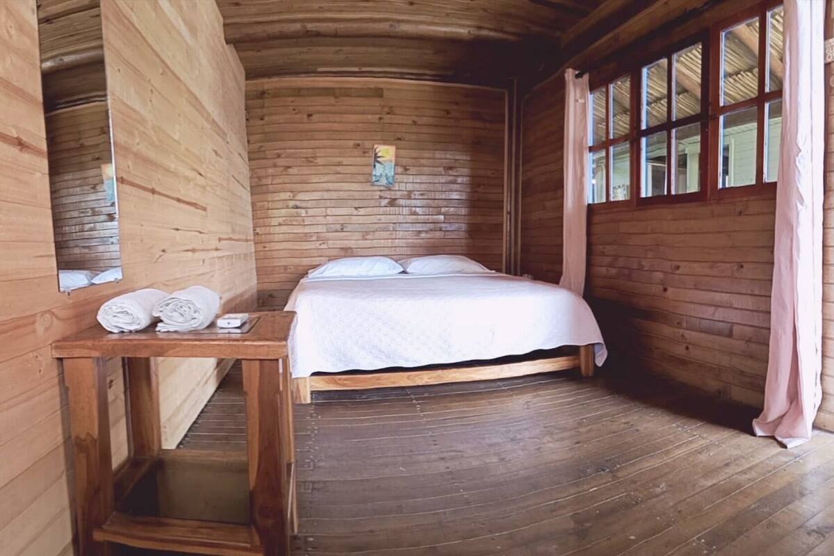 Une chambre dans un chalet en bois, avec un lit, une petite table sur laquelle sont posées des serviettes et une fenêtre avec des rideaux.