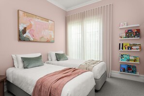 A bedroom with two beds, a large window with sheer curtains, a bookshelf with children's books, and a colorful abstract painting on the wall.