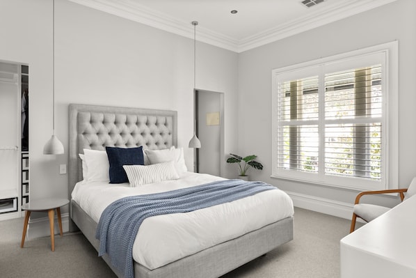 A bedroom with a bed, bedside lamps, a window with blinds, a plant, and a chair.