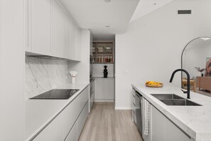 A modern kitchen with white cabinetry, a black induction cooktop, a wooden dining table, and a fruit bowl.