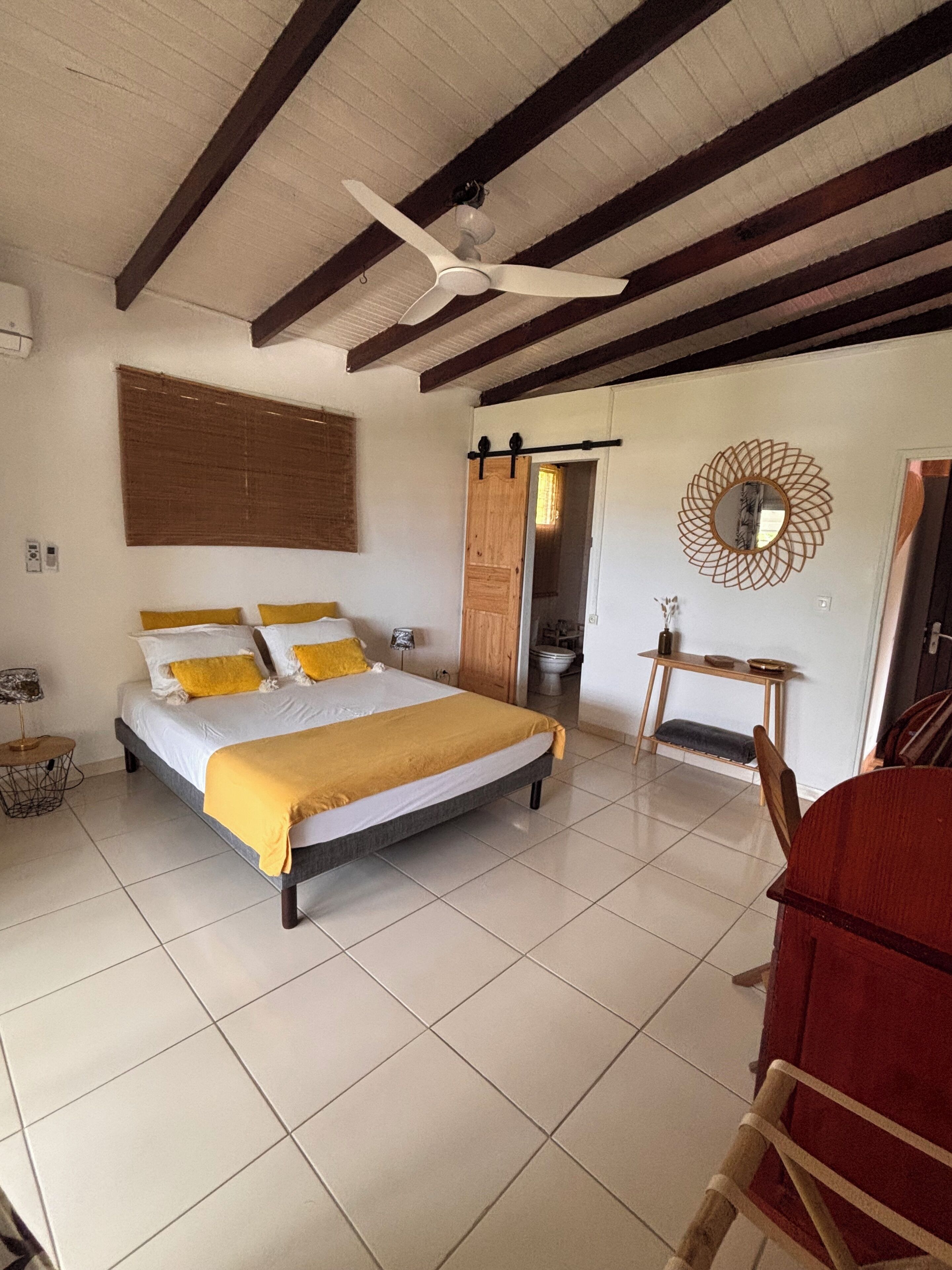 A bedroom with a bed, a ceiling fan, a wooden door, a mirror, and a small table.