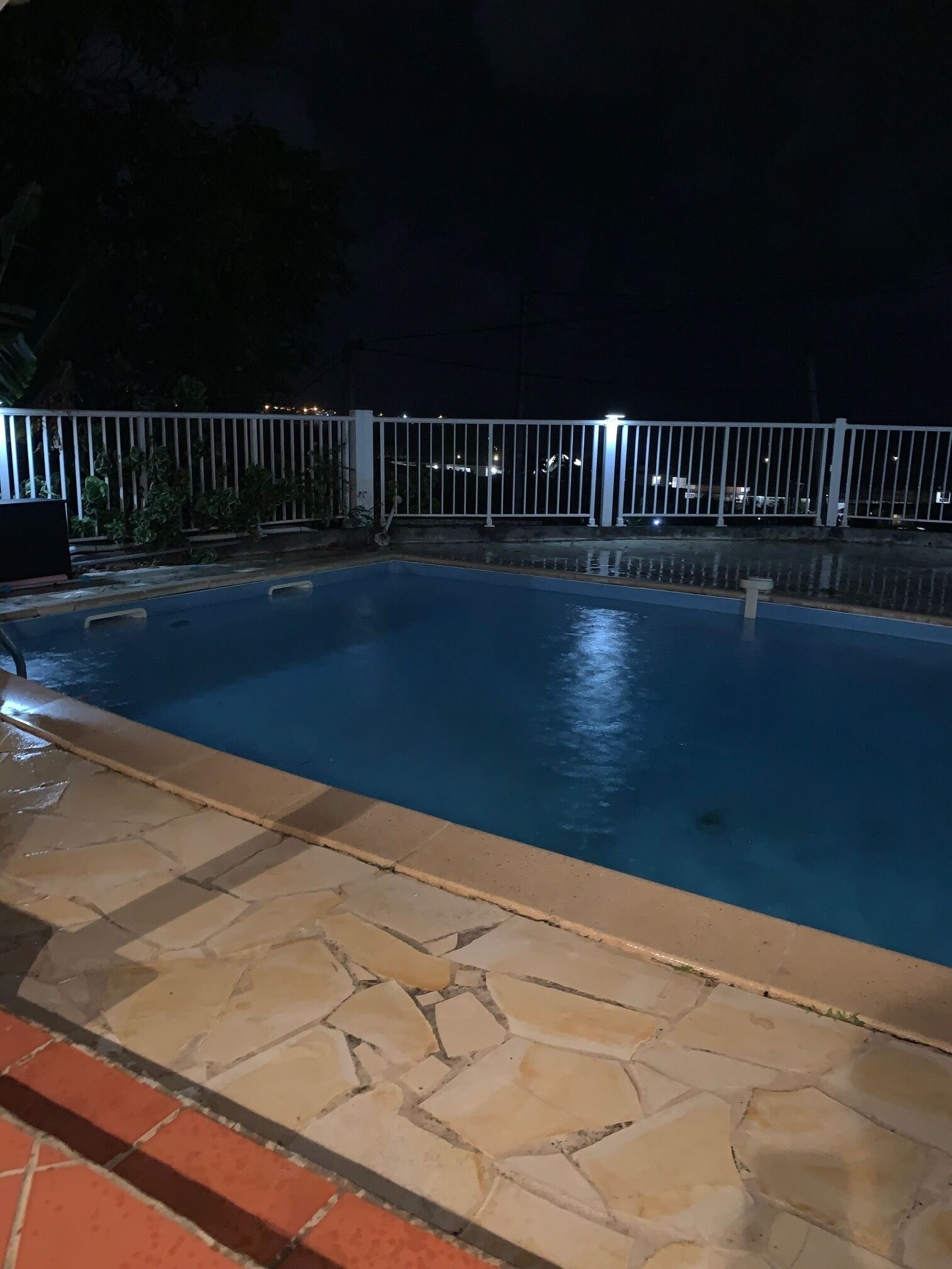 A nighttime swimming pool with a stone-tiled deck and a white metal fence.
