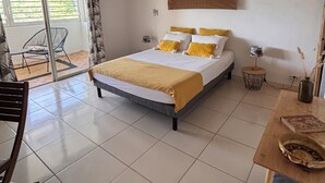 A bedroom with a bed, a ceiling fan, an air conditioning unit, a window with blinds, a chair, and a small table with a vase. - Notre Vie Là (Le Robert)