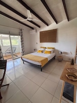 Un dormitorio con cama, ventilador de techo, equipo de aire acondicionado, ventana con persianas, una silla y una mesita con un florero.
