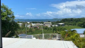A view from a balcony with a metal roof overlooking a coastal area with buildings, greenery, and a body of water. - Notre Vie Là (Le Robert)