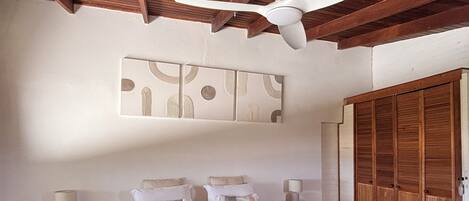 Une chambre à coucher avec un lit, des poutres en bois au plafond, un ventilateur de plafond, une œuvre d’art accrochée au mur, une table de chevet avec une lampe et une armoire en bois.