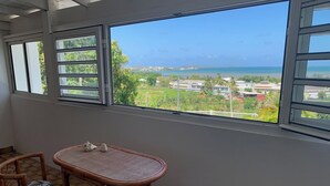 A balcony with a view of a beach and houses. There is a wicker table and two wicker chairs with cushions. - Notre Vie Là (Le Robert)