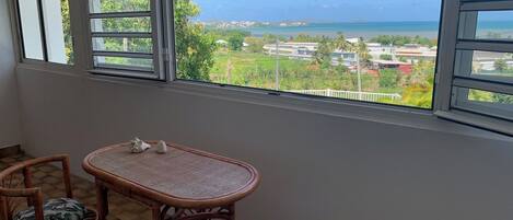 A balcony with a view of a beach and houses. There is a wicker table and two wicker chairs with cushions.