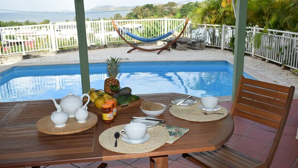 A wooden table set for breakfast with white teapots, cups, and a bowl of fruit, overlooking a swimming pool and a white fence. - Notre Vie Là (Le Robert)