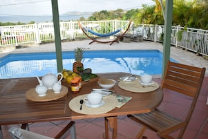 Una mesa de madera preparada para el desayuno, con teteras blancas, tazas y un cuenco de fruta, con vistas a una piscina y una valla blanca.