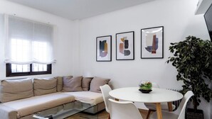 A modern living room with a beige sofa, a glass coffee table, and a round dining table with white chairs. There are abstract paintings on the wall and a large green plant in the corner. - Spacious, modern, and central 2-bedroom apartment with 1 bathroom in Madrid (Madrid)