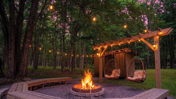 Un foyer circulaire entouré de bancs en bois, une pergola en bois décorée de guirlandes lumineuses et des fauteuils suspendus, le tout situé dans une zone boisée.