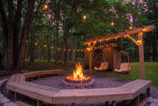 A circular fire pit surrounded by wooden benches, a wooden pergola with string lights, and hanging chairs in a forested area.